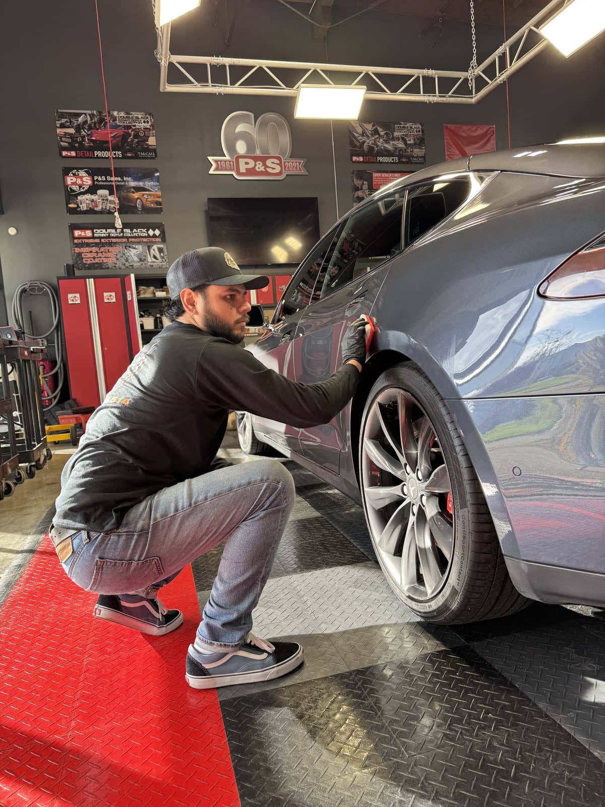 Detailer crouched at the rear wheel of a grey car, working the lower paint.